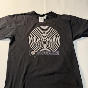 May include: Black t-shirt featuring a large white and black graphic design. The design includes concentric circles and the Adidas logo. The word "ADIDAS" is printed below the graphic in a stylized font. The shirt has a crew neck and short sleeves.