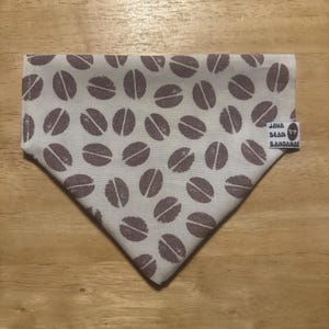May include: A triangular dog bandana with a repeating pattern of brown coffee beans on a cream background. The bandana has a tag that reads "Java Bean Bandanas".