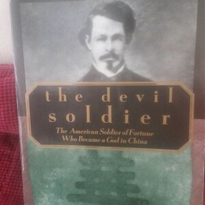 Op de afbeelding: Boekomslag met een zwart-witfoto van een man met een snor en baard. De titel van het boek is "The Devil Soldier: The American Soldier of Fortune Who Became a God in China" door Caleb Carr.