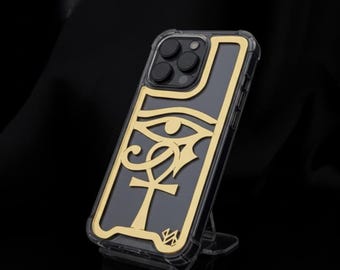 Pode incluir: Capa de telefone transparente com um design dourado do Olho de Hórus egípcio. A capa tem um fundo preto e uma moldura dourada. A capa do telefone está apoiada em um suporte transparente.