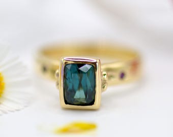 May include: A gold ring featuring a rectangular, teal-colored gemstone. The ring has a gold band with small decorative elements and additional gemstones. The design is elegant and suitable for various occasions.