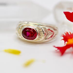 May include: A silver ring featuring a deep red, oval gemstone. The ring has an engraved pattern around the stone. The background is soft white, with a blurred red flower in the corner.
