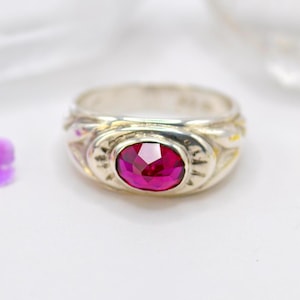 Op de afbeelding: Een zilveren ring met een ovale, gefacetteerde, robijnrode edelsteen. De ring heeft een decoratieve band met geëtste details rond de edelsteen en langs de zijkanten. De ring is geplaatst tegen een zachte, witte achtergrond.
