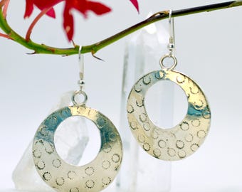 May include: Silver-toned hoop earrings with a textured surface and small dot patterns. The earrings are circular with a large open center. A red flower and a clear crystal are in the background.