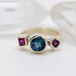 May include: A silver ring featuring a central round blue gemstone flanked by two square, magenta-colored gemstones. The ring has a simple band and a modern design, perfect for everyday wear.