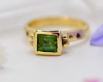 May include: A gold ring with a square, green gemstone at the center. The ring has a gold band with small, decorative accents. The gemstone is a vibrant green color, set in a gold bezel.