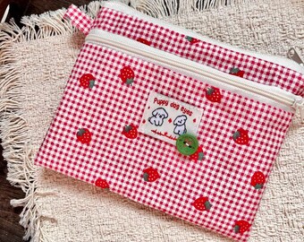 May include: A red and white gingham patterned pouch with strawberry designs. The pouch has a white zipper and a small label that says "Puppy dog eyes". A green button is sewn on the pouch.