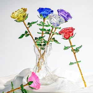 以下が含まれることがあります： 透明なガラスの花瓶に、カラフルなガラスのバラのブーケが入っています。バラは黄色、青、透明、紫、赤です。各バラは、緑の葉が付いた金色の茎を持っています。ピンクのバラが花瓶の前にあります。