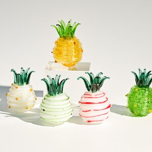 Peut inclure: Plusieurs ananas décoratifs en verre de différentes couleurs. Les ananas ont des sommets verts et sont jaunes, blancs, verts et rouges. Un ananas est sur une petite plateforme blanche.