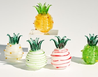 May include: A collection of decorative glass pineapples in various colors. The pineapples have green leafy tops and are yellow, white, green, and red. One pineapple is elevated on a small white platform.