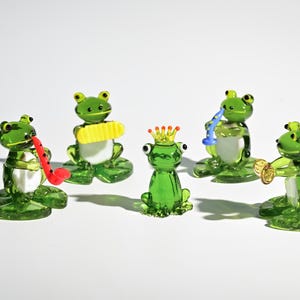 以下が含まれることがあります： ガラス製のカエル5体のコレクション。それぞれが楽器を演奏しています。緑色のカエルは白いお腹をしており、サックス、クラリネット、トランペット、王冠が描かれています。