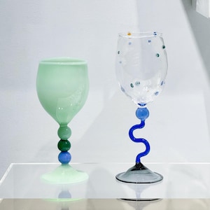 May include: Two decorative wine glasses. One is mint green with a stem of stacked blue and green spheres. The other is clear with blue and green dots and a blue, wavy stem. Both glasses have a unique, artistic design.