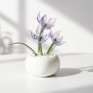 May include: A decorative glass sculpture featuring three translucent purple flowers with green stems and yellow centers, planted in a small, round white ceramic pot filled with white pebbles. The piece displays delicate, hand-crafted glass petals.