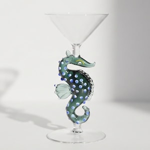 Puede incluir: Una copa de martini de cristal transparente con un tallo en forma de caballito de mar verde oscuro. El caballito de mar tiene lunares azul claro, una pequeña aleta y un detalle de ojo amarillo. La copa tiene un cuenco cónico y base circular.