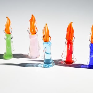 Puede incluir: Cinco figuras de velas de vidrio de colores. Cada vela tiene una base de diferente color: verde, rosa, azul, rojo y azul. Las llamas son naranjas y amarillas. Las velas están dispuestas sobre una superficie blanca.