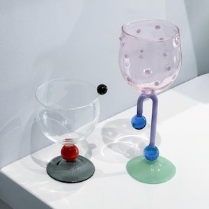 May include: Two decorative wine glasses. One is clear with a red sphere and a gray base. The other is pink with a purple stem, blue spheres, and a green base. The pink glass has small, raised dots.