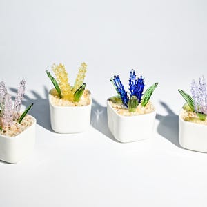 May include: Four small, white square planters each hold a different colored glass flower arrangement. The flowers are in shades of lavender, yellow, and blue, with green glass leaves. The planters sit on a white surface.