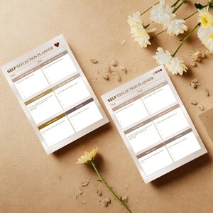 May include: Two self-reflection planners with a heart icon and the text "Self Reflection Planner". Each planner has sections for positive affirmations, self-compassion, and gratitude. The planners are on a tan surface with flowers.