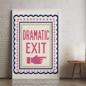 May include: A rectangular cross-stitch wall art piece featuring a cream-coloured fabric background. The text DRAMATIC EXIT is stitched in pink, accompanied by a pink pointing hand icon. It is framed by decorative blue, pink, and green borders.