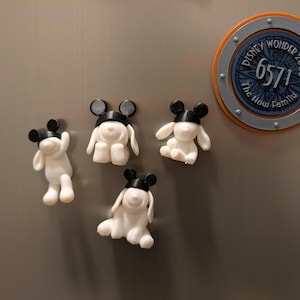 May include: White 3D-printed cartoon character figurines with black ears and hats. The figurines are in different poses. A circular magnet with the text "Disney Wonder 2025" and "The How Family" is also visible.