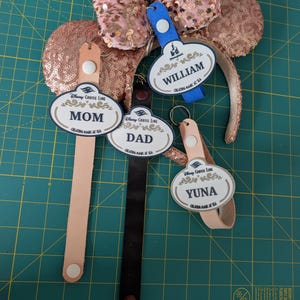 Peut inclure: Un serre-tête oreilles de souris à paillettes or rose avec des étiquettes nominatives personnalisées. Les étiquettes indiquent "MOM", "DAD", "WILLIAM" et "YUNA". Les étiquettes sont attachées à des lanières en cuir. Le logo Disney Cruise Line est sur les étiquettes.