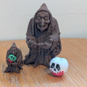 Puede incluir: Tres figuras de brujas marrones y una manzana blanca con diseño de calavera. La figura de bruja más grande está de pie, mientras que la más pequeña sostiene una manzana verde y roja. La manzana con la calavera tiene goteo rojo desde abajo.