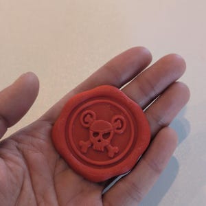 Könnte beinhalten: Ein runder, roter Wachssiegel mit einem Totenkopf- und gekreuzten Knochen-Design, umgeben von einem dekorativen Rand. Der Schädel hat mausohrähnliche Formen. Das Siegel wird in einer Hand gehalten.