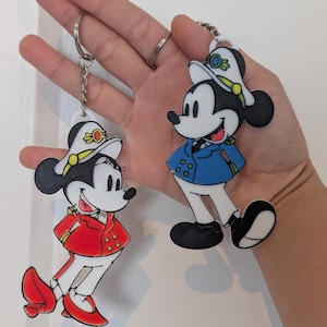 Può includere: Due portachiavi di Topolino. Un Topolino indossa una giacca rossa e l'altro una giacca blu. Entrambi hanno cappelli bianchi con dettagli dorati. I portachiavi sono attaccati a portachiavi argentati.