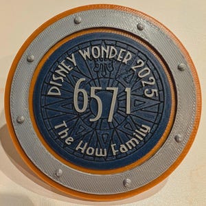 Peut inclure: Un objet décoratif rond multicouche avec le texte "DISNEY WONDER 2025" et "6571" dans une police stylisée. Il présente un centre bleu, un anneau central gris argenté et un anneau extérieur orange. Le texte "The How Family" est également présent.
