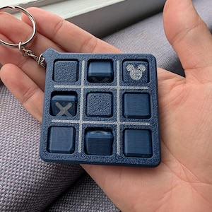 May include: A navy blue tic-tac-toe keyring with a silver ring. The game board features a grid of nine squares, with an "X" and a Mickey Mouse head design. The keyring is held in a hand.