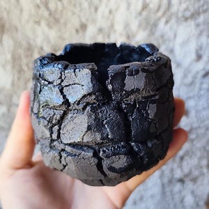 Könnte beinhalten: Ein kleines, rundes, schwarzes Keramikgefäß mit einer stark texturierten, rissigen und unebenen Oberfläche. Die Außenseite weist tiefe Spalten und erhabene, gezackte Segmente auf, die ein raues, steinartiges Erscheinungsbild erzeugen.