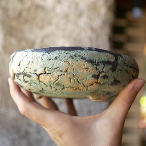 May include: A round ceramic bowl with a textured, crackled surface featuring a blend of light green and earthy tan tones. The rim is dark, and the base includes small, short feet. The bowl has a weathered, organic appearance with visible fissures.