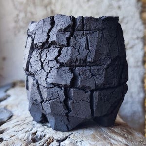 Op de afbeelding: Een kleine, donker houtskoolkleurige keramische plantenbak met een zwaar getextureerd, gebarsten en gespleten buitenoppervlak. De pot heeft een cilindrische vorm met een ruwe, onregelmatige rand en een basis die rust op korte, blokvormige voetjes.