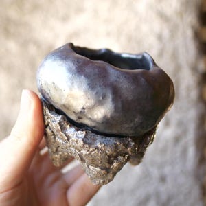 Op de afbeelding: Een kleine keramische kom met een gladde, donker-metalen afwerking aan de bovenzijde en een ruwe, getextureerde, bronskleurige basis met drie korte pootjes. De kom heeft een onregelmatige, organische rand en een diepe, ronde binnenkant.