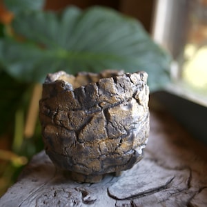 Op de afbeelding: Een kleine, ronde keramische pot met een sterk getextureerd, ruw buitenoppervlak. De pot heeft een donker antracietgrijze basiskleur met metallic gouden accenten op de verhoogde, onregelmatige randen, wat een steenachtig uiterlijk creëert.