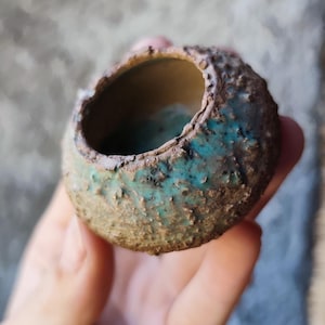 May include: A small, round ceramic vessel with a textured, rough exterior surface. The pot features a mottled color palette of earthy brown and vibrant turquoise blue glaze, with a smooth, lighter-toned interior visible through the circular opening.