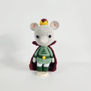 May include: A handmade crochet mouse doll wearing a green tunic, a burgundy cape, and a yellow crown. The doll has gray fur, pink ears, and black eyes. It stands on a small, round, beige base.