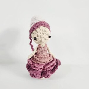 May include: A handmade crochet doll with a light beige head, black eyes, and a purple and white hat. The doll wears a layered pink dress. The doll's arms and legs are also crocheted. The doll is a charming handmade item.