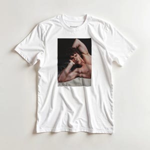 May include: A white short-sleeved t-shirt featuring a rectangular photographic print on the chest. The print displays a person with curly hair resting their head on one hand, with their arm raised, captured in a high-contrast, dimly lit setting.