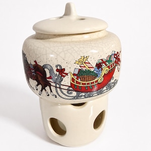 May include: A cream-coloured ceramic oil warmer with a crackle glaze finish. The upper section features a colourful illustration of a brown horse pulling a red sleigh filled with wrapped gifts. The base has three circular cutouts for ventilation.