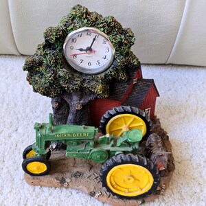 May include: A decorative table clock featuring a green John Deere tractor, a red barn, and a large tree. The clock face is white with black numerals and hands, set into the tree. The base is textured brown, and a small dog figure stands nearby.
