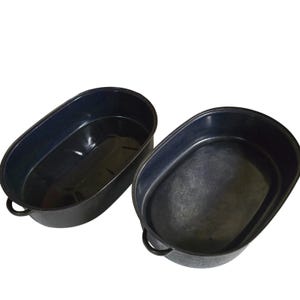 May include: Two oval-shaped roasting pans with a dark navy blue enamel finish. Each pan features a smooth interior surface and two small, rounded metal handles on the sides. The pans are open at the top and have a deep, basin-like structure.