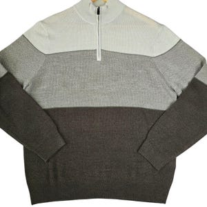 May include: A long-sleeve quarter-zip sweater featuring a color-blocked design with a light cream upper section, a medium gray middle section, and a dark charcoal lower section. The garment has a ribbed mock neck collar and a central zipper closure.
