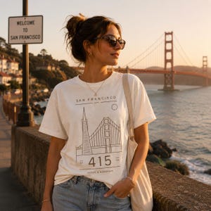 May include: Cream-colored t-shirt with a San Francisco graphic featuring the Golden Gate Bridge and the Transamerica Pyramid. The shirt includes the text "SAN FRANCISCO" and "415". The person is wearing sunglasses and standing near the Golden Gate Bridge.