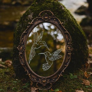 May include: An oval-shaped mirror with a decorative, ornate bronze frame. The mirror's surface reflects a blurred outdoor scene and features two white line-drawn hummingbirds. The mirror is resting on a moss-covered rock in a natural setting.