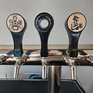 Pode incluir: Três torneiras de cerveja com alavancas pretas e bicos prateados. As torneiras têm tampas circulares de madeira com ilustrações de homens segurando canecas de cerveja. Uma torneira diz "Lager" e a outra "Morzen".