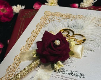 May include: A burgundy rose pen with a pearl center, gold accents, and a white feather, resting on a wedding certificate. The pen is embellished with gold detailing and a cream-colored ribbon. The certificate has Arabic script and signatures.