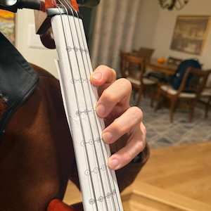 Puede incluir: Primer plano de un violín con una tabla de digitación adherida. El violín es de color marrón rojizo, con clavijas y cuerdas negras. Una mano está colocada en el diapasón, con una tabla de papel que muestra las posiciones de las notas. El fondo está borroso.