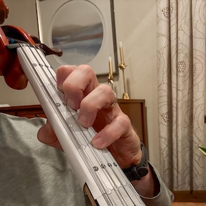 May include: A close-up of a violin with a white fretboard overlay. The overlay has black lines and markings. A person's fingers are pressing down on the strings. The violin is brown and the background is blurred.