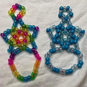 Könnte beinhalten: Zwei Perlenfiguren, eine in Regenbogenfarben und die andere in Blautönen. Die Figuren haben einen Ring am Boden und eine sternartige Form oben, verbunden durch Perlenstränge. Die Perlen sind durchscheinend und undurchsichtig.